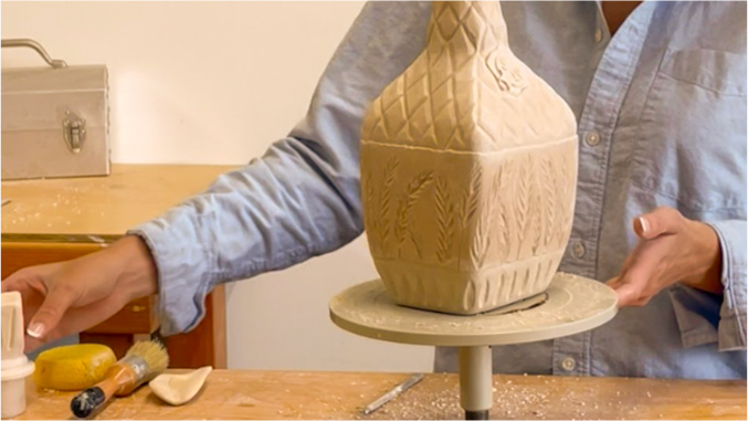 A potter shapes a decorative clay vase on a spinning wheel, tools and materials scattered on a wooden table nearby.