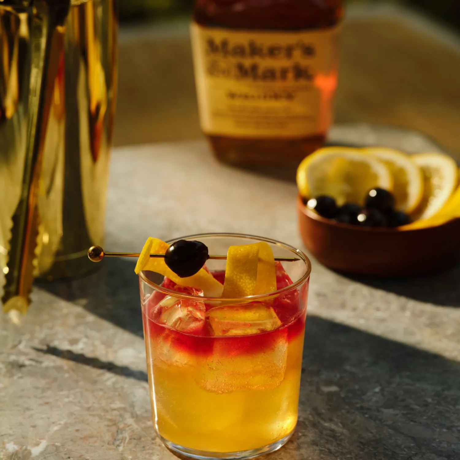 A glass of whiskey cocktail with ice, garnished with a lemon peel and cherry, sits on a table. A whiskey bottle, bowl of lemon slices, and a golden shaker are in the background. The setting conveys a warm, elegant mood.