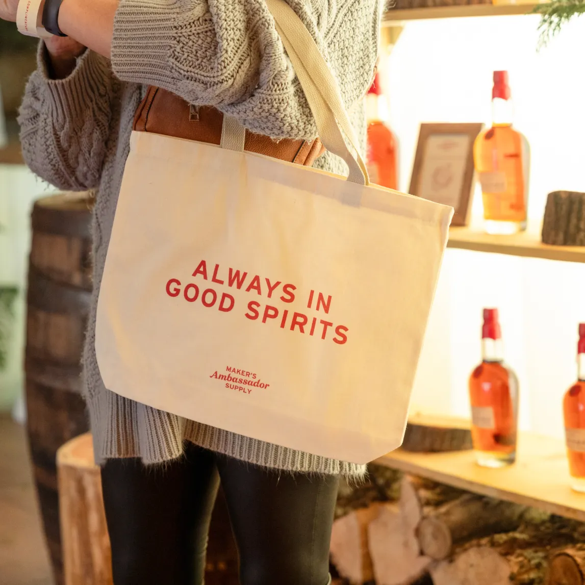 A person holds a tote bag reading "ALWAYS IN GOOD SPIRITS" in front of a display of whiskey bottles on a wooden shelf.