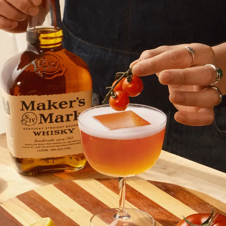 A hand adds cherry tomatoes to a cocktail garnished with a clear ice cube, alongside a bottle of Maker's Mark bourbon on a wooden table.