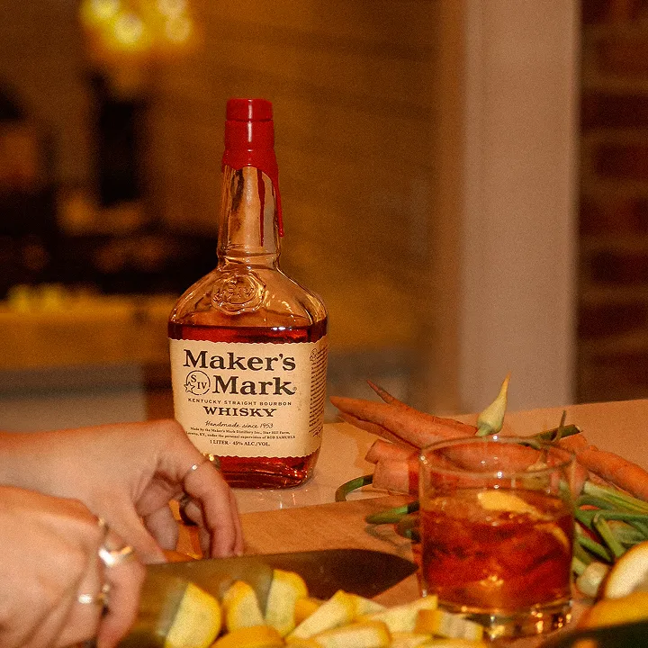 A bottle of Maker's Mark bourbon on a table next to a glass with a drink, surrounded by chopped fruits and fresh carrots.