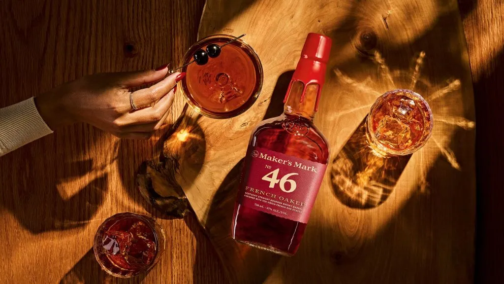 A hand with a cocktail garnished with cherries beside a bottle of Maker's Mark 46 and two glasses on a wooden surface.