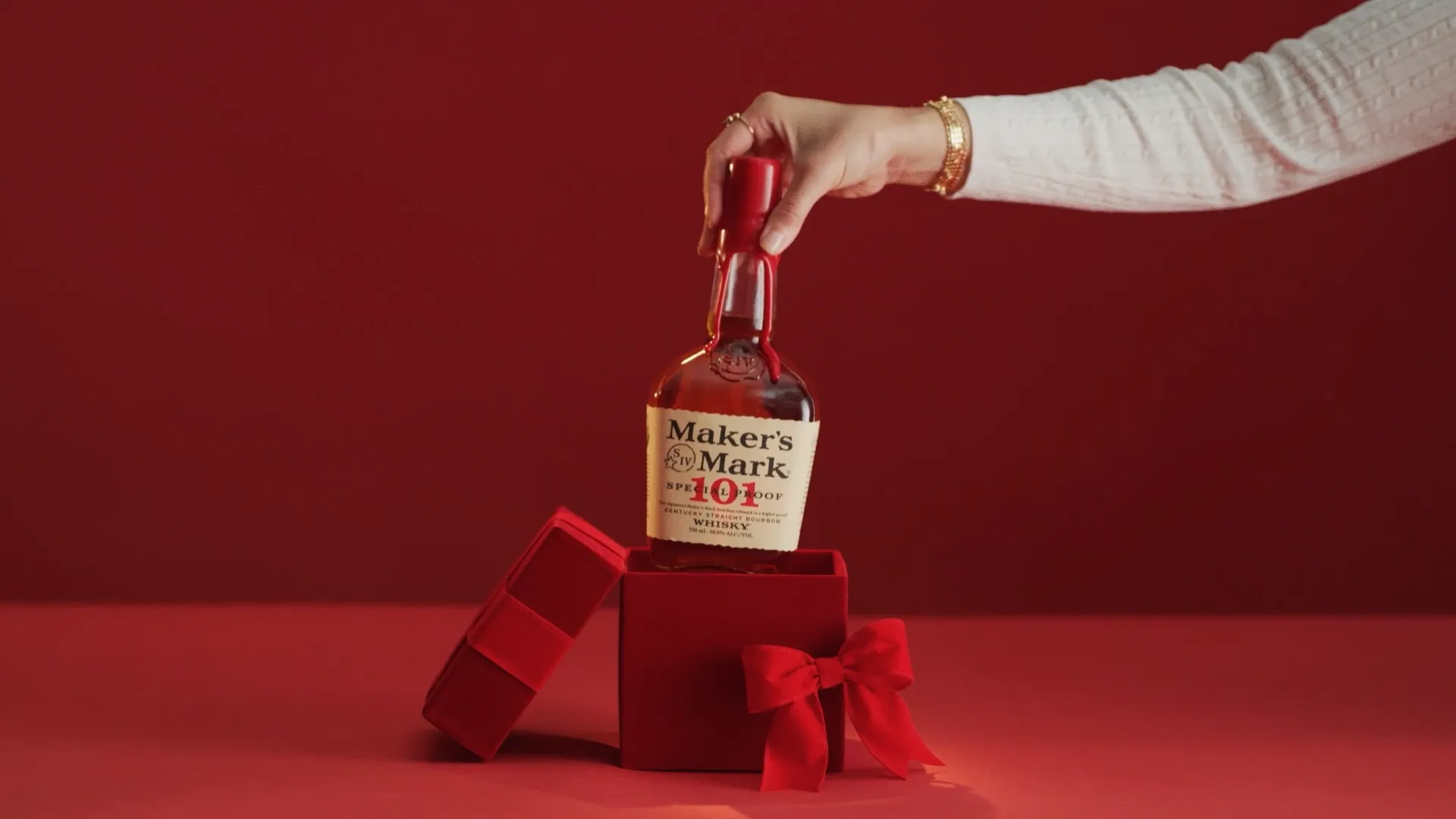 A hand reaches into a red box to lift a bottle of Maker's Mark 101 Bourbon, set against a matching red background.
