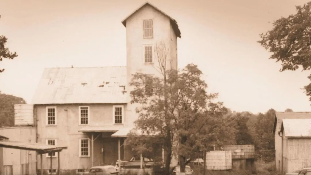 Historic grain mill with a tall tower, surrounded by trees and outbuildings, in a sepia-toned photograph. Classic rural setting.