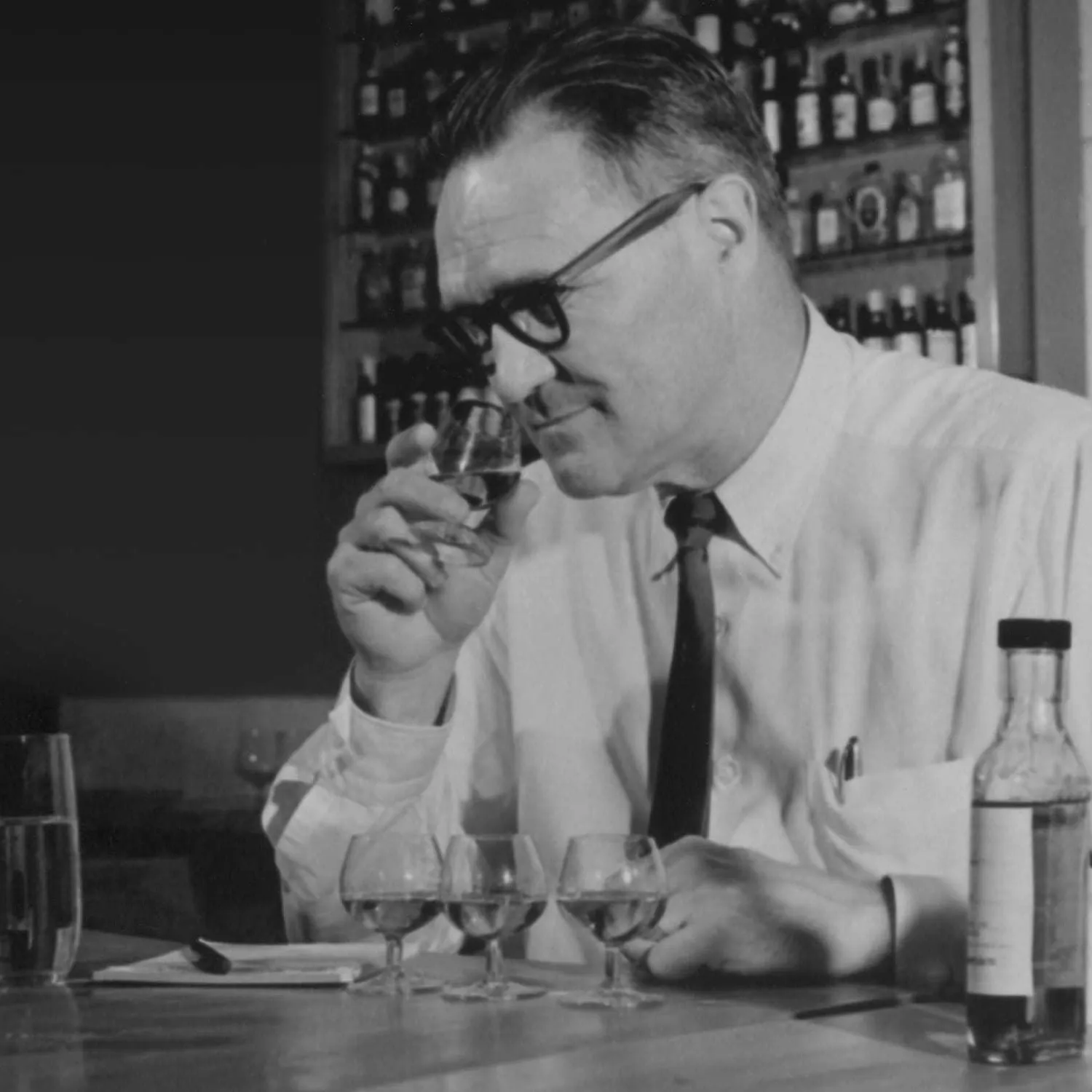 A man in a white shirt and tie samples liquids from small glasses, surrounded by bottles, in a vintage bar setting.